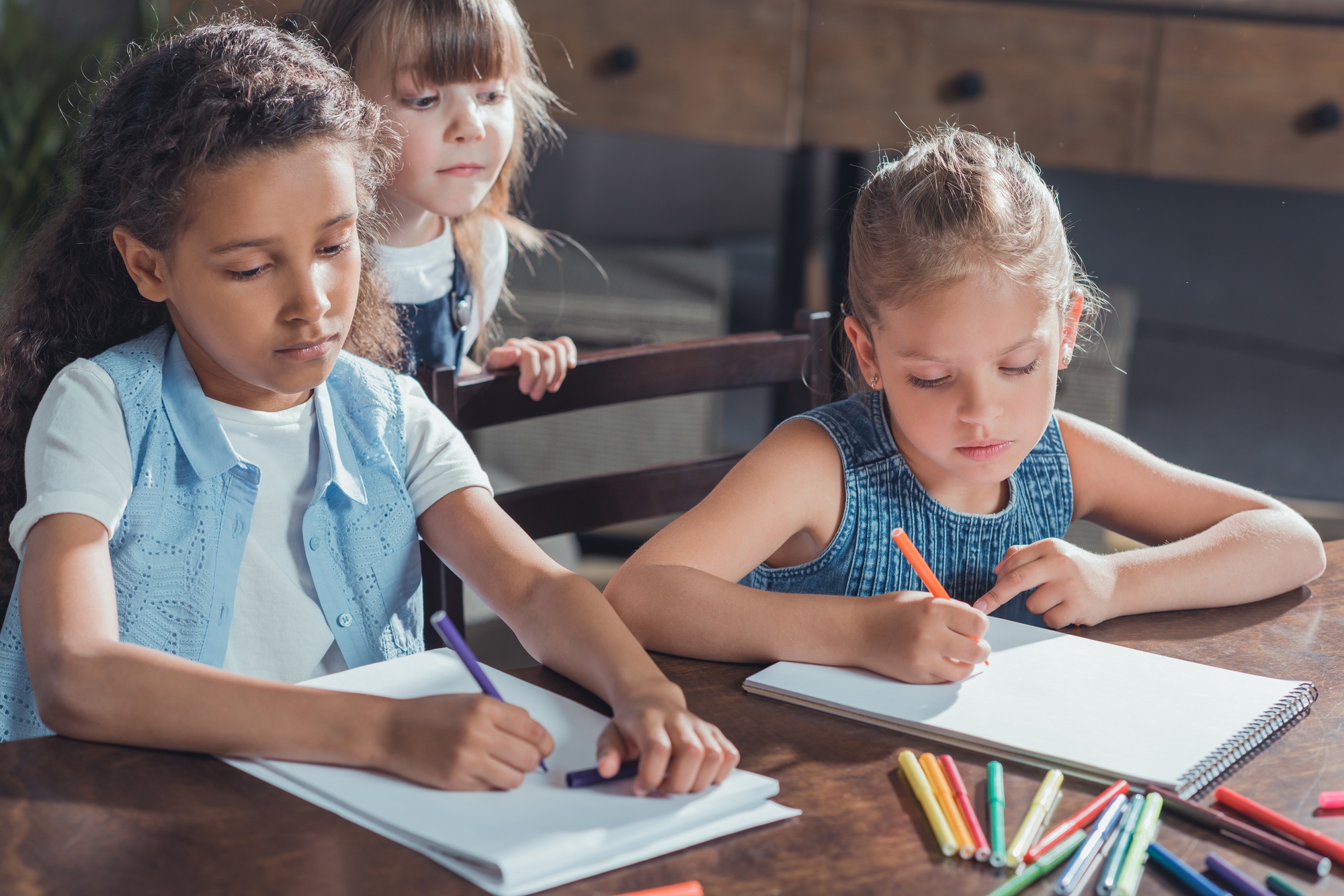 little girl looking how friends drawing pictures with colorful markers at home