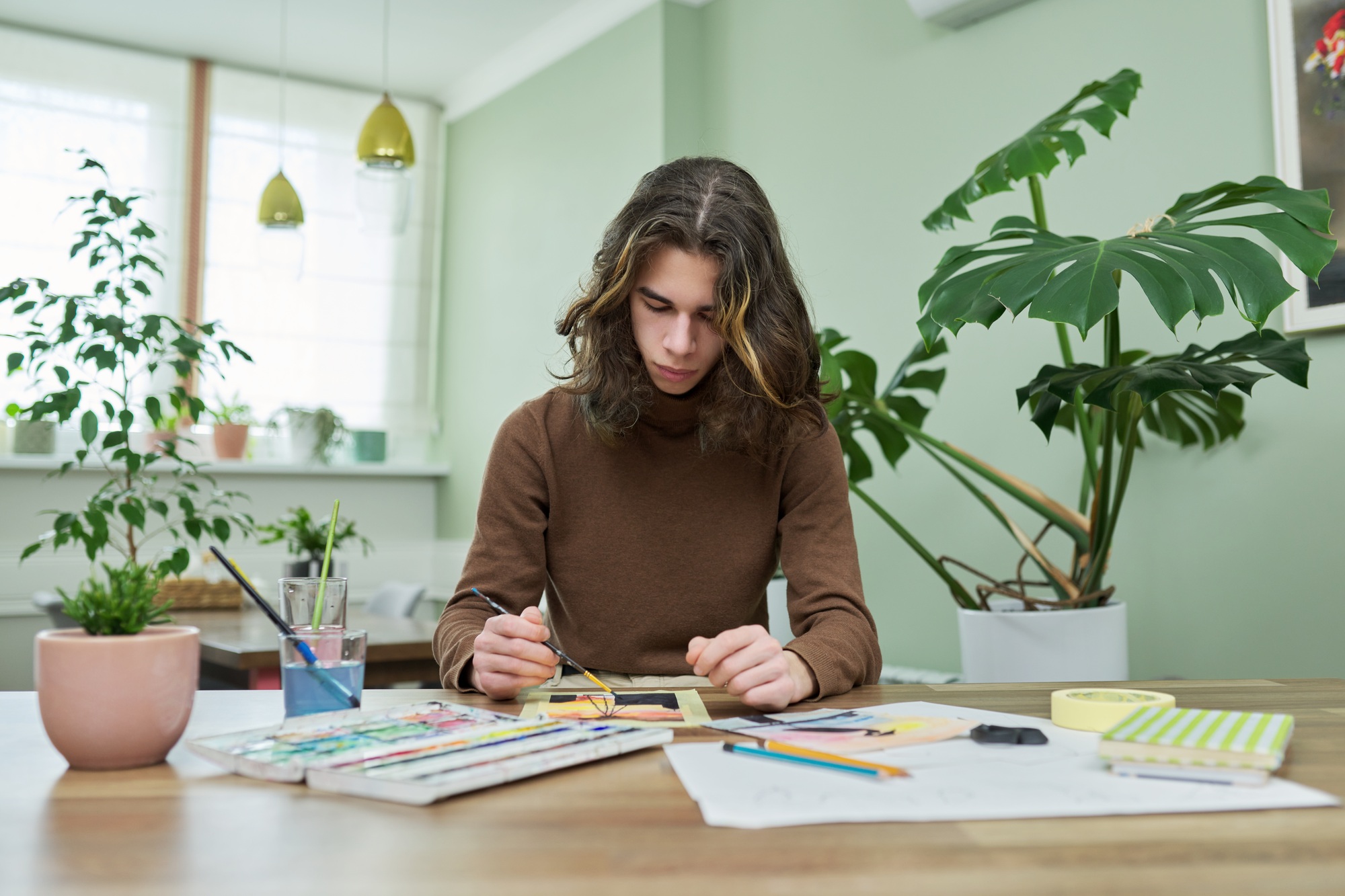 Talented creative guy teenager painting with watercolors, sitting at the table at home